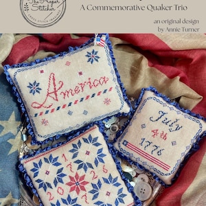 May include: A patriotic cross-stitch trio featuring the word "America", "July 4th 1776", and a starburst design. Each piece is framed with blue ribbon and adorned with red, white, and blue cross-stitch details. The design is by Annie Turner.