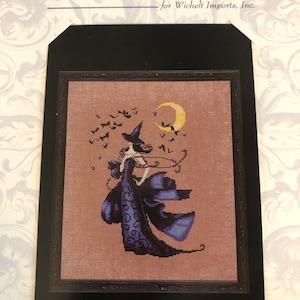 May include: Cross-stitch design by Nora Corbett, featuring a witch in a flowing purple gown, a crescent moon, and bats. The artwork is framed in black and set against a tan background. The text "Design by Nora Corbett" is at the top.