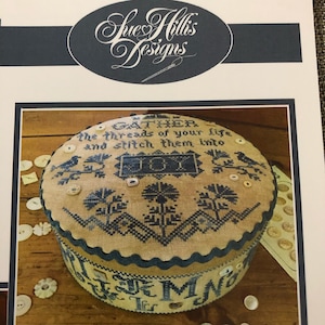 May include: A round, beige fabric box with blue embroidery. The top of the box features the text "Gather the threads of your life and stitch them into JOY". The bottom of the box has the text "Life is the most precious gift we have".