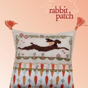 May include: A white pillow with a brown rabbit and carrots embroidered on it. The pillow has a green border and a white background with a carrot pattern. The pillow has two orange tassels on the corners. The pillow is labeled "rabbit patch" and "Cecelia Lewis".