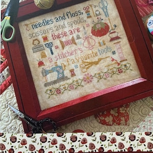 May include: A framed cross-stitch pattern with a red frame and a white background. The pattern depicts various sewing tools, including needles, thread, scissors, spools, and a sewing machine. The text "needles and floss, scissors and spools, these are a stitcher's favourite tools" is stitched in the center of the pattern. The title "A Stitcher's Tools" is printed below the frame, along with the designer's name, "Lori Pengelly".
