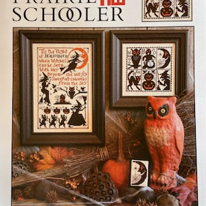May include: A cover for a cross-stitch pattern book with a Halloween theme. The cover displays framed cross-stitch samplers with witches, cats, and pumpkins. The title "Sweeping Cobwebs" is shown below the images. An orange owl statue and a small pumpkin are also visible.