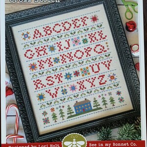 May include: A framed cross-stitch sampler featuring the text "Home Town Holiday Sampler" and a colourful alphabet. The design incorporates festive motifs such as trees and a house. The frame is dark with an ornate pattern.