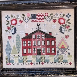 May include: Framed cross-stitch depicting a red building, an American flag, and the text "AMERICA THE BEAUTIFUL." The design incorporates floral and berry patterns, a weathervane, and stylized trees. The artwork is set within a black frame, evoking a patriotic theme.