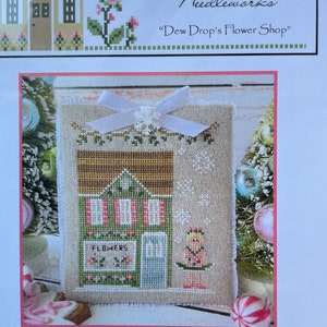 May include: Cross stitch pattern for a Nutcracker Village shop called "Dew Drop's Flower Shop". The pattern features a small building with a green awning and a sign that says "Flowers". A ballerina stands in front of the shop.