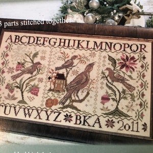 May include: A cross-stitch sampler with a cream background featuring a bird motif, a small house, and a poinsettia. The sampler is framed in a dark brown wood frame and includes the alphabet, the letters "BKA", and the year "2011".