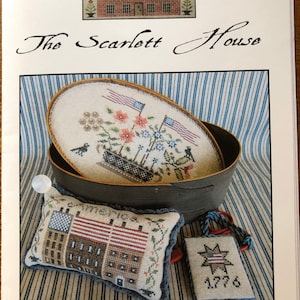 May include: A patriotic shaker box set with a red, white, and blue theme. The set includes a round box with a lid, a pillow, and a small tag. The box features a floral design with American flags. The pillow has a house design with American flags. The tag has a star design with the year 1776.
