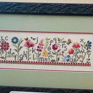 May include: A cross-stitch embroidery of the alphabet with flowers and butterflies. The letters are in a white font with a red outline. The flowers are in shades of pink, red, yellow, and blue. The butterflies are yellow and black.