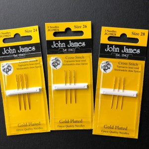 May include: Three packages of gold-plated cross stitch needles in sizes 24, 26, and 28. Each package has a yellow background with the John James logo and the text "Cross Stitch Tapisserie bout rond Sticknadeln ohne Spitze" and "Gold Plated Finest Quality Needles".