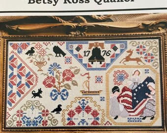 May include: A framed cross-stitch sampler with the title "Betsy Ross Quaker". The sampler features a variety of patriotic symbols, including a bell, a ship, a deer, and a flag. The sampler is stitched in a variety of colors, including red, white, blue, and gold.