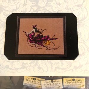May include: Cross-stitch pattern featuring a witch riding a broom, with a black frame and the text "Design by Nora Corbett." Below are three packages of glass seed beads for needlework projects.