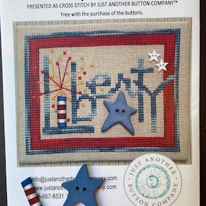 May include: A cross-stitch pattern titled "Liberty Star" with a patriotic theme. The design features the word "Liberty" in blue, red, and gold, with star-shaped buttons and a firework design. The package includes additional star-shaped buttons and a red, white, and blue button.