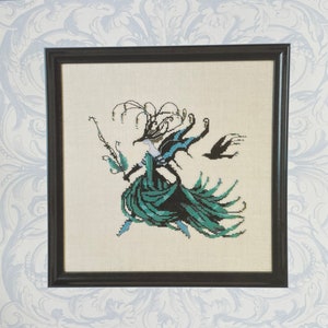 May include: A cross-stitch pattern featuring a whimsical, stylized woman in a green dress and black hat. The pattern is titled "Masqued Mischief" and is designed by Nora Corbett for Weichel Imports, Inc. The pattern number is NC295.