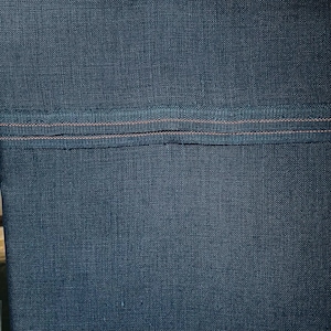 May include: Close-up of a section of dark blue fabric with a textured weave. A horizontal band of darker blue fabric is sewn across the center, with two parallel lines of orange stitching.
