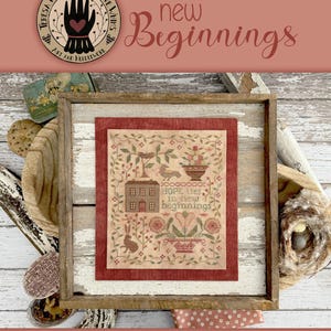 May include: A cross-stitch pattern featuring a house, flowers, and a bird with the text "Hope lies to new beginnings". The pattern is framed in a wooden frame and is set against a rustic background.