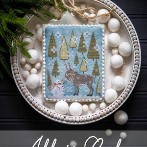 May include: A cross-stitch Christmas ornament with a winter scene featuring a reindeer, snowman, and trees. The ornament is displayed on a white tray with decorative white balls and greenery. The text "All is Calm" is at the bottom.