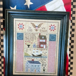 May include: Framed sampler with a black wooden frame. The sampler features patriotic imagery, including an eagle, American flags, and text that reads "In God We Trust." The sampler is a cross-stitch design with a vintage aesthetic.