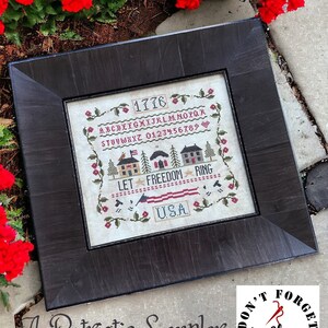 May include: A framed cross-stitch sampler with the text "Let Freedom Ring" and "1776" in red, white, and blue. The sampler also includes a small American flag, houses, and the text "USA". The caption below the sampler reads "A Patriotic Sampler Snack" and "Don't Forget to Floss" with a needle and thread graphic. The text "Finally A Farmgirl" is below the graphic.