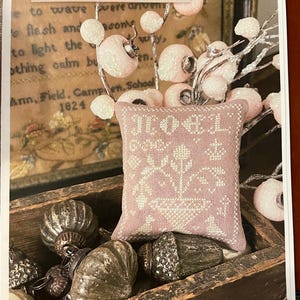 May include: A small, pink embroidered pillow with the word "NOEL" and a floral design. The pillow is in a wooden box with vintage-style ornaments. The text "NOEL TUFFET by Shakespeare's Peddler" is at the top.