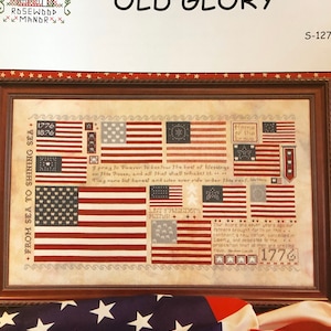 May include: Framed cross-stitch artwork titled "Old Glory" featuring various American flags. The design includes the text "From Sea to Shining Sea" and patriotic quotes. The artwork is in a wooden frame.