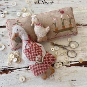 May include: A handcrafted, fabric goose and duck, part of the "Animal Cracker Series + Olive+". The goose has a red and white patterned body, and the duck has a red bow. A decorative pillow with floral embroidery and the text "Animal Cracker Series + Olive+" is also present.