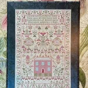 May include: Framed sampler titled "The Checkerboard House Sampler" by Eliza Gagin, 1842. The cross-stitch design features a red and blue house, floral and bird motifs, and a border with text. The frame is dark with a textured appearance.