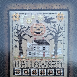 May include: A cross-stitch design featuring a Halloween theme. The design includes a jack-o'-lantern, a house, ghosts, and bare trees. The word "HALLOWEEN" is stitched across the bottom. The text "Halloween Saltbox" is at the top.