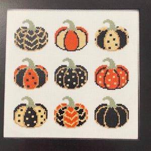 May include: Nine cross-stitched pumpkins in a square arrangement. Each pumpkin has a green stem and is decorated with various patterns and colors, including black, orange, and cream. The pumpkins are on a white background.