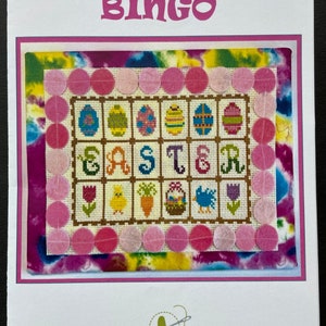 May include: A cross-stitch pattern for an Easter Bingo game. The pattern features a brown background with a pink and white border. The centre of the pattern features the word "Easter" in colourful letters, surrounded by images of eggs, chicks, carrots, and a basket of eggs. The pattern is surrounded by a border of pink felt circles.