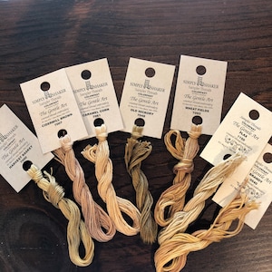 May include: Close-up of eight spools of embroidery floss in various shades of brown and beige. Each spool has a label with the text "Simply Shaker", "Sampler Threads", "Colorfast", "The Gentle Art", and a number. The labels are arranged in a row from left to right.