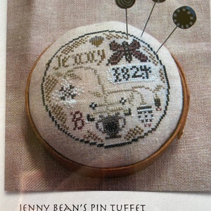 May include: A round, cream-colored pincushion with a brown wooden frame. The pincushion is embroidered with a butterfly, flowers, and the text "Jenny 1824". Three decorative pins are stuck into the pincushion.