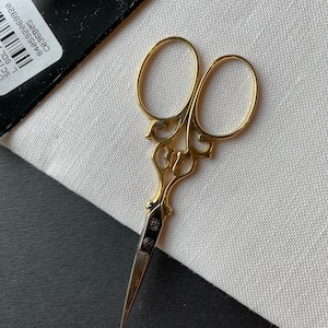 May include: Gold-colored embroidery scissors with ornate handles and sharp blades. The scissors are resting on a white fabric, with a black surface in the background. The scissors have a decorative design.