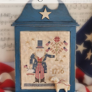 May include: A blue wooden framed patriotic cross-stitch design with the text "One Flag, One Land!" and "1776". The design features a figure, flags, and a tree with flags. A small sheep figurine is in front of the frame.