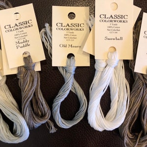 May include: Five spools of embroidery floss in various shades of gray and white. The spools are labeled with the brand name "Classic Colorworks" and the color names "Muddy Puddle", "Old Money", "Snowball", and "Stepping Stones".
