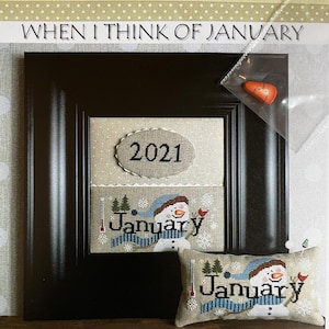 May include: A framed cross-stitch design with the text "WHEN I THINK OF JANUARY". The design features a snowman with a blue scarf and hat, a red bird, and the word "January". A small pillow with the same design is also visible.