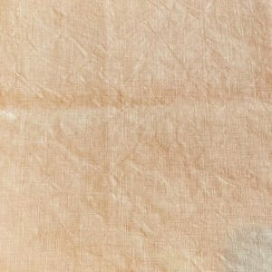 May include: Close-up of a textured, light peach-colored fabric. The fabric has a woven appearance with visible threads and subtle variations in tone, creating a soft, natural look. The material is slightly wrinkled.