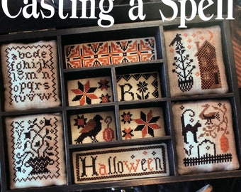 May include: A wooden box with nine compartments, each containing a small embroidered piece. The pieces feature various Halloween-themed designs, including a house, a cat, a raven, and a pumpkin. The box is labeled "Casting a Spell" and "Halloween".