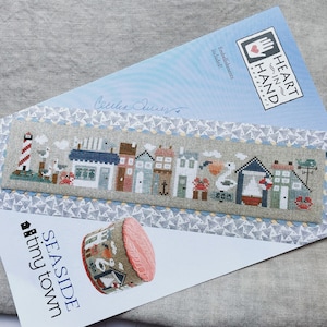 May include: A cross-stitch pattern for a seaside town with a lighthouse, houses, and a seagull. The pattern is called "Seashore Tiny Town" and is from the Heart in Hand collection. Embellishments are included.