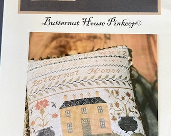 May include: A cross-stitch cushion featuring a yellow house, two black cauldrons, and the text "Butternut House" within a brown frame. The cushion is displayed on a white background with a brown fringe.