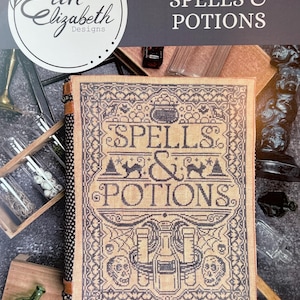May include: A cross-stitch pattern book titled "SPELLS & POTIONS" with a beige background and dark blue stitching. The cover features the text "SPELLS & POTIONS" and illustrations of a cauldron, cats, bottles, and skulls. The book is bound with a patterned spine.