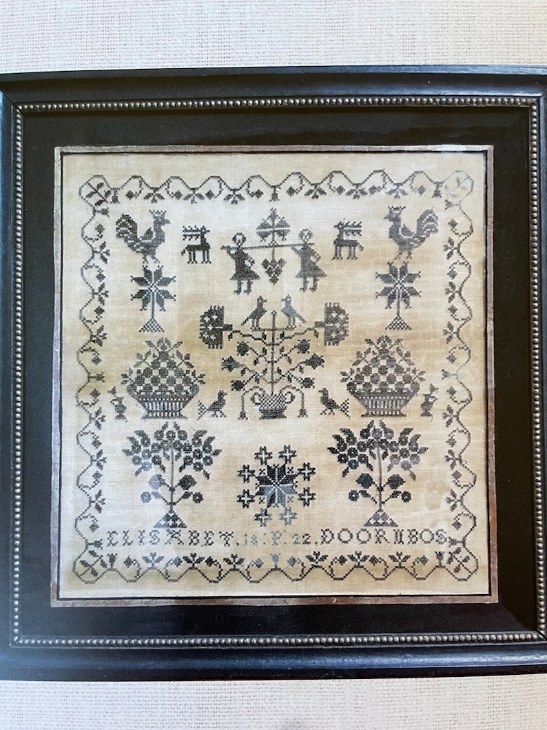 Może przedstawiać: Czarno-biały haftowany sampler z motywem kwiatowym, dwoma koszami kwiat&oacute;w, kogutkiem, jeleniem i dwiema postaciami trzymającymi laski. Sampler jest oprawiony w czarną ramę z ozdobnym obramowaniem z koralik&oacute;w. Na dole haftowany jest tekst "ELISABET. 18. 22. DOORIBOS".