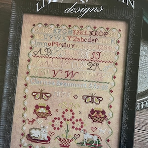 May include: A framed cross-stitch sampler with a vintage aesthetic. The sampler features a floral design with butterflies, baskets, and a cat and lamb. The text "Clarissa Beaumont - 1875" is at the bottom of the sampler.