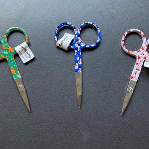 May include: Three pairs of small scissors with colorful patterned handles. The scissors have a green, blue, and white and red design. The handles are labeled "Embroidery Scissors".