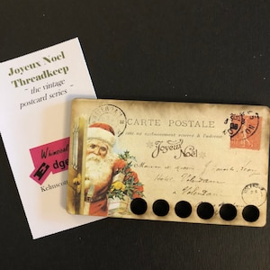 May include: A vintage postcard featuring a Santa Claus illustration with a red suit, white beard, and a bag of gifts. The postcard is a thread keeper with eight holes for storing thread. The postcard has the text "CARTE POSTALE" and "Joyeux Noel" printed on it.