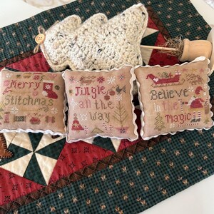 May include: Three small, square, beige Christmas pillows with white frilled edges, each embroidered with festive phrases and images. A crocheted Christmas tree and a wooden ornament are in the background. The pillows are on a green and red patterned fabric.