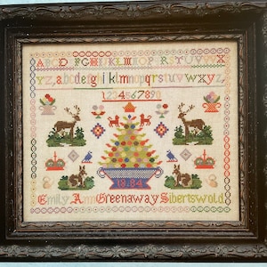 May include: A framed cross-stitch sampler with a colorful Christmas tree design. The sampler includes the alphabet, numbers, and the text "Emily Ann Greenaway Sibertswold 1831-1894".