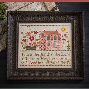 May include: A framed cross-stitch sampler with a red brick house, flowers, and birds. The text reads "This is the day that the Lord hath made. We will rejoice and be Glad in it." Below the frame, the text reads "This is the Day! Original Designs by Paulette Stewart."