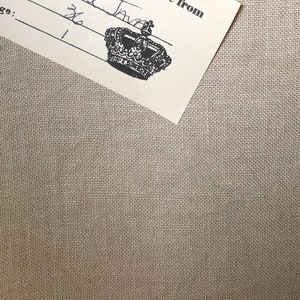 May include: A piece of light beige, woven fabric with a visible texture. Attached to the fabric is a white rectangular tag featuring a black crown icon and printed text that reads: color: Old Mill Java, at: 36, ge: 1, and reproductions, hand-dyed fabric from.