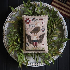 May include: A rectangular, beige decorative pillow with a cross-stitch design featuring an eagle, the year 1776, and patriotic stars. The pillow is surrounded by a white beaded tray and green foliage, with a small American flag in the background.