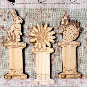 May include: Three wooden thread bobbins with a bunny, daisy, and strawberry design. The bobbins are engraved with the text "Heartstring Samplery".